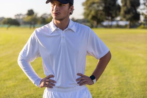 Male athlete standing on sunlit field wearing white sportswear, cap and smartwatch