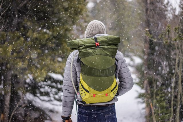 Backpacker wearing grey jacket and beanie hiking through snow-covered forest trail. Ideal for travel blog, adventure group promotions, outdoor gear advertisements, or articles about winter hiking and nature exploration.