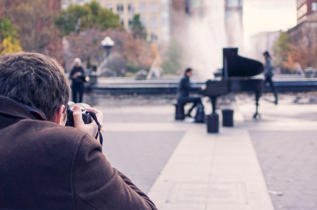 This scene features a man photographing a pianist playing a grand piano in an urban park. The background is blurred, showing a fountain, trees with fall colors, and other people. This visual is perfect for themes related to hobbies, street performance, urban life, and fall activities.