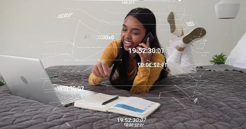 Young woman working remotely on laptop while talking on phone with planner on bed, smiling