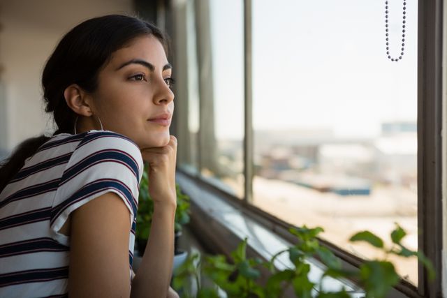 Young woman in a striped shirt gazing thoughtfully out of an office window, surrounded by indoor plants. Ideal for use in articles or advertisements about workplace wellness, mental health, daydreaming, or modern office environments. Can also be used for lifestyle blogs or inspirational content.
