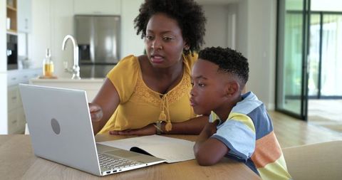 African american mother guiding son through online learning on laptop in modern kitchen