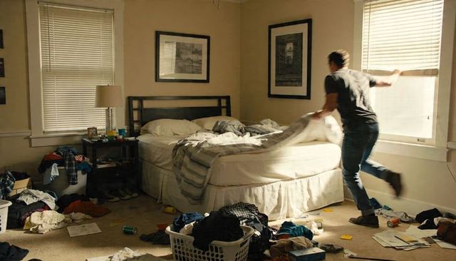 Young man pulling blind while making bed in messy sunlit bedroom, morning chore motion