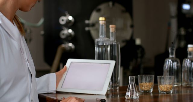 Woman in white coat working in laboratory, using tablet to collect data. Glass bottles, flasks, and beakers containing liquids and grains visible on table. Ideal for depicting scientific research, modern technology in lab settings, or females in STEM fields.