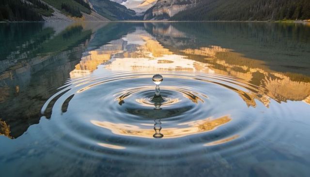 Single water droplet creating concentric ripples on alpine lake reflecting sunlit cliffs