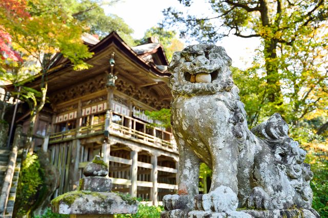 Traditional Japanese shrine featuring a stone guardian statue amidst vibrant autumn foliage. Ideal for travel brochures, cultural presentations, and websites promoting Japanese heritage and tourism. Highlights themes of spirituality, nature, and tranquility.