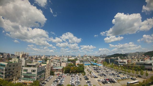 A vibrant cityscape showcasing various buildings under a bright clear blue sky dotted with fluffy white clouds. The image also displays an aerial view of the city's infrastructure including residential and commercial buildings as well as a parking lot filled with cars. Ideal for urban planning websites, travel brochures, and city guides.