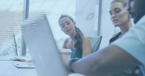 Woman leading collaborative team meeting in glass-walled office with laptops and coffee