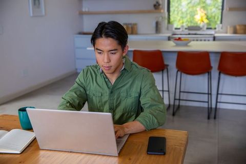 Asian man working on laptop at home dining table bright modern kitchen remote work