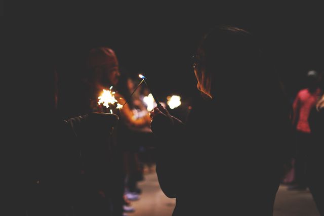 Silhouetted people holding sparklers against a dark background during a nighttime celebration. The image conveys a festive and joyful atmosphere, suitable for use in promotional materials, event invitations, or holiday-themed advertisements.