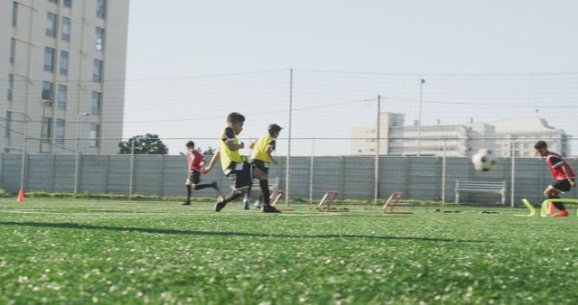 Young boys in yellow bibs running on a green soccer field during a practice session. Urban buildings in background indicating a city setting. Photo is ideal for promoting youth sports programs, physical activity, teamwork values, or urban sports facilities.