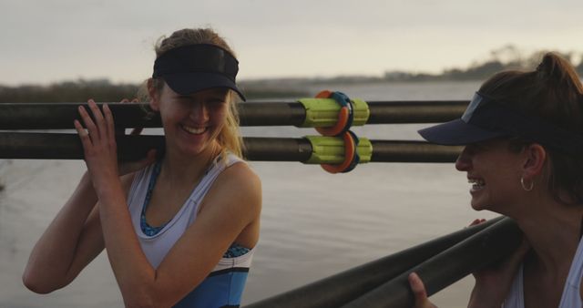 Two female rowers are seen laughing and chatting as they carry their oars by the water. They appear to be enjoying their time together, showcasing themes of teamwork, camaraderie, and outdoor fitness. This image is suitable for illustrating sportsmanship, female empowerment in sports, and recreational activities.