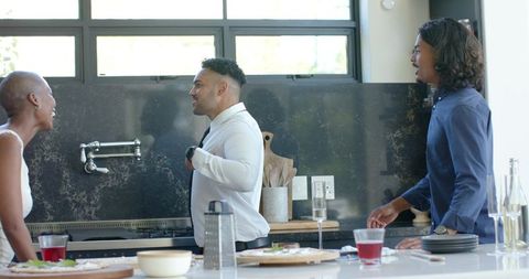 Man adjusting tie at kitchen island while friends laughing during casual home gathering