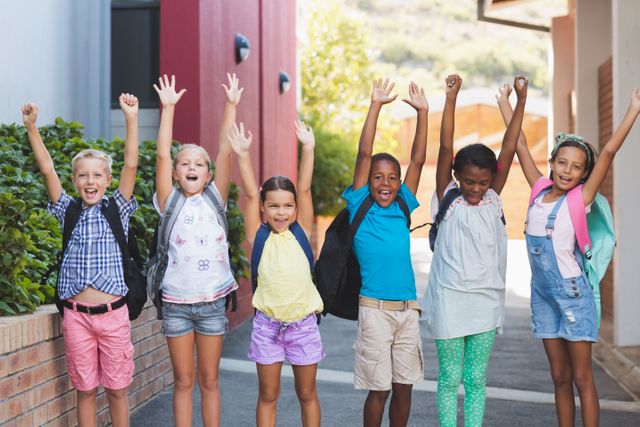 Group of cheerful children standing in a row with their hands raised, showing excitement and happiness. They are wearing casual clothes and backpacks, indicating they are at school. This image can be used for educational materials, back-to-school promotions, advertisements for children's products, or articles about childhood and education.