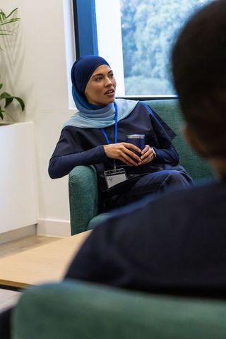 Woman wearing blue hijab sitting in office lounge holding mug and talking with colleague
