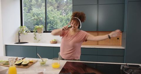 Joyful woman dancing while making guacamole in modern kitchen with headphones enjoying juice