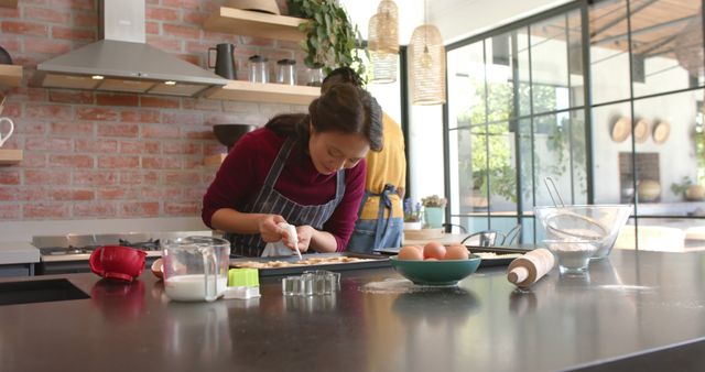 Woman decorating freshly baked cookies in a modern kitchen with bright and natural light, suggesting a relaxed home baking setting. Perfect for use in content related to baking, recipes, cooking tutorials, home life, family activities, and culinary blogs.