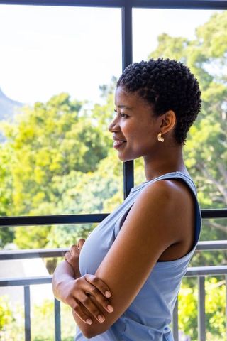 Contemplative african woman standing on balcony crossing arms and looking through window