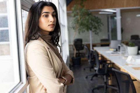 Indian professional woman leaning at window in modern office, contemplative leader portrait
