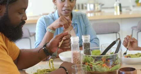 African family sharing cozy kitchen dinner, eating pasta and tossing fresh salad