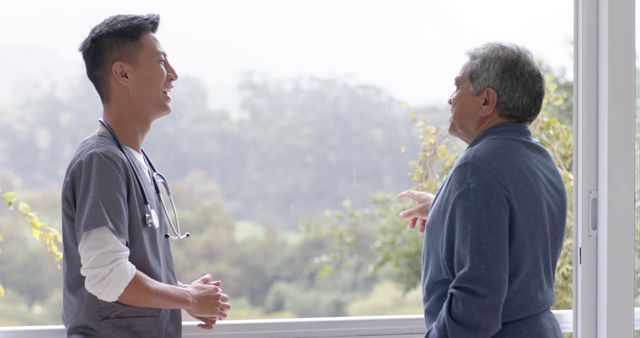 Healthcare professional engaging in conversation with elderly male patient by window. Perfect for promoting doctor-patient relationships, elderly care services, healthcare discussions, wellness programs.