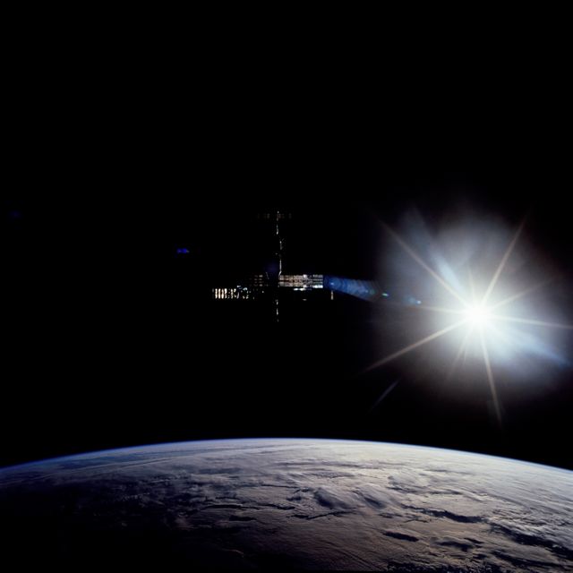 Image showing Space Shuttle Discovery approaching the Mir Space Station against the backdrop of the blackness of space, with parts of Mir illuminated and Earth visible below. Useful for illustrating historic space missions, collaboration between space agencies, space exploration themes, and educational materials on 1990s space history.