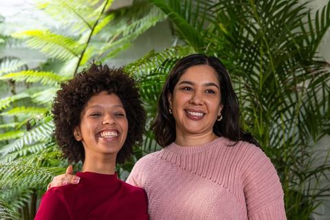 Smiling diverse women standing in greenhouse touching shoulder among lush tropical ferns
