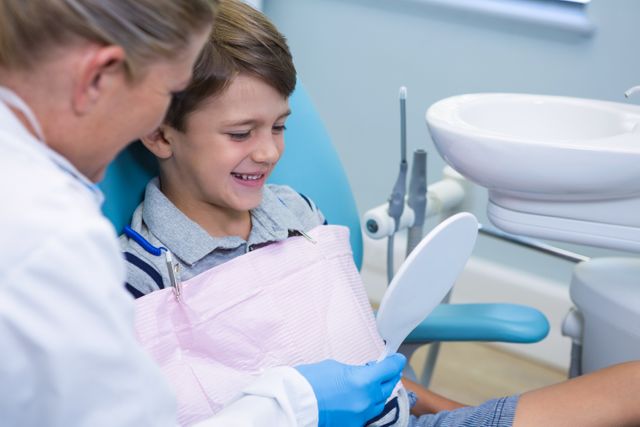 Dentist showing mirror to smiling boy at dental clinic. Ideal for use in articles or advertisements about pediatric dental care, oral health, dental checkups, and healthcare services for children.