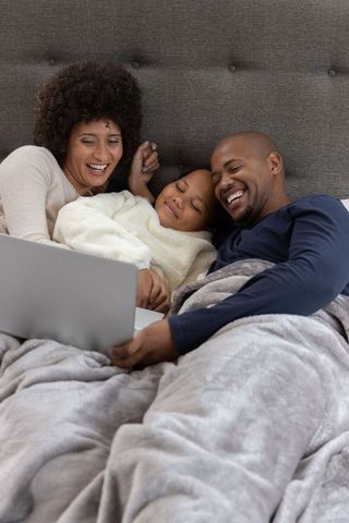 Diverse family laughing and watching laptop together in cozy bedroom