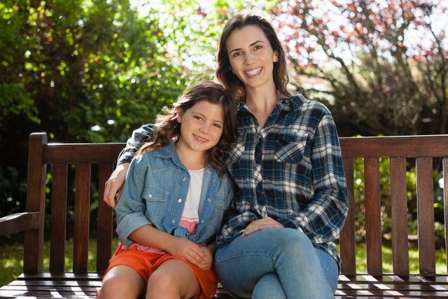 This image depicts a smiling mother and daughter sitting on a wooden bench in a backyard. The mother is wearing a plaid shirt and jeans, while the daughter is dressed in a denim jacket and orange shorts. The background features lush greenery and blooming trees, suggesting a sunny day. This image can be used for family-oriented content, advertisements, blogs about parenting, outdoor activities, and lifestyle articles.