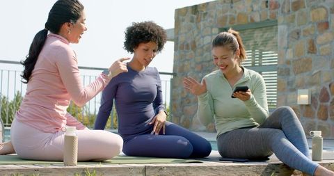 Diverse women practicing yoga and chatting on rooftop patio with phones and water bottles
