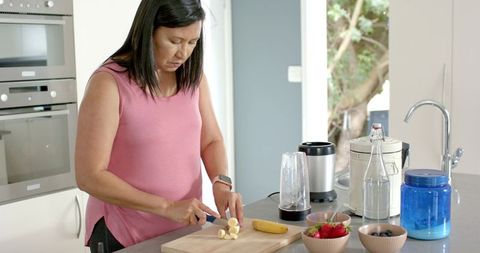 Woman slicing banana on wooden board preparing healthy smoothie at modern kitchen island