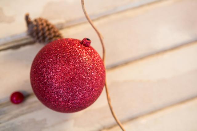 Red glitter Christmas bauble lying on a wooden plank with a pine cone in the background. Ideal for holiday greeting cards, festive decorations, and seasonal marketing materials.