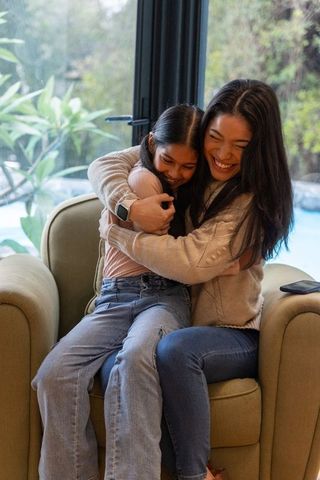 Mother hugging daughter on armchair by sliding glass doors, smiling cozy family moment