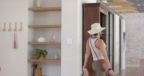 Woman wearing wide-brimmed hat walking through bright minimalist hallway holding brochure
