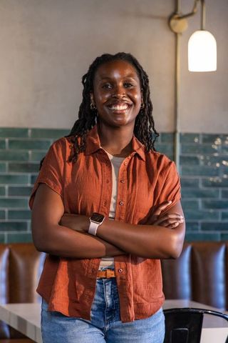 Confident african woman smiling with arms crossed in urban cafe booth wearing rust shirt