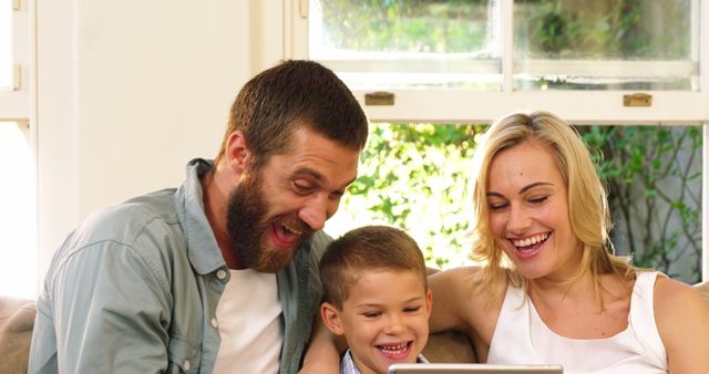 Happy family sitting on sofa in living room, parents and son enjoying time together using digital tablet. Perfect for advertisements on family products, technology for kids, online services, educational content, and home interiors.