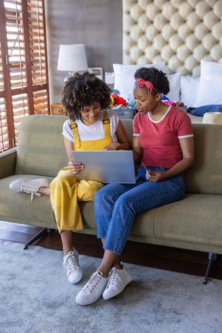Two friends sharing laptop on sofa in cozy bedroom planning trip and bookings