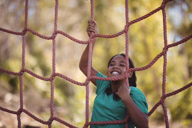 Young girl climbing a net during an obstacle course, smiling and enjoying the challenge. Ideal for use in advertisements for summer camps, outdoor activities, fitness programs for children, and educational materials promoting physical activity and adventure.