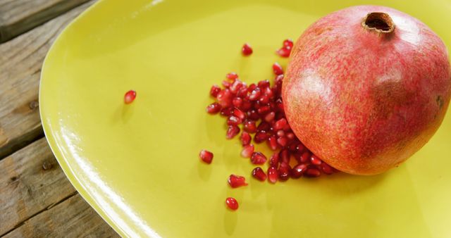 Whole pomegranate and scattered seeds on a bright yellow plate resting on wooden surface. Ideal for concepts related to healthy eating, fresh produce, and nutrition. Perfect for use in cookbooks, health blogs, and food-related websites.