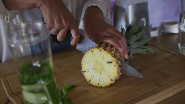 Person slicing fresh pineapple on wooden board emphasizes home-cooking themes, as well as detox and smoothie ingredient preparation. Great for illustrating concepts of health, wellness, organic food preparation, and detox recipes.