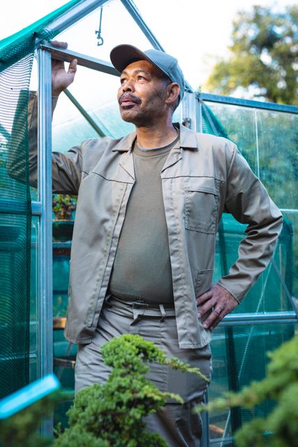 Senior male gardener standing confidently in a greenhouse, surrounded by plants. Ideal for use in articles or advertisements about gardening, horticulture, independent businesses, or senior activities. Can also be used for content related to plant care, greenhouse management, and sustainable agriculture.
