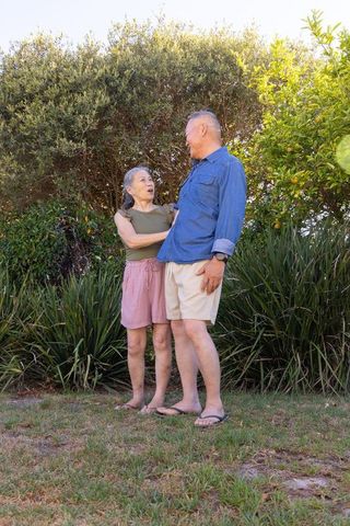 Sunlit senior couple laughing and embracing while standing in backyard garden with lush greenery