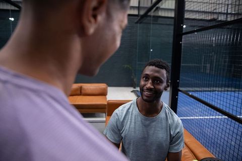 Diverse men relaxing and chatting on orange leather couch beside indoor paddle court