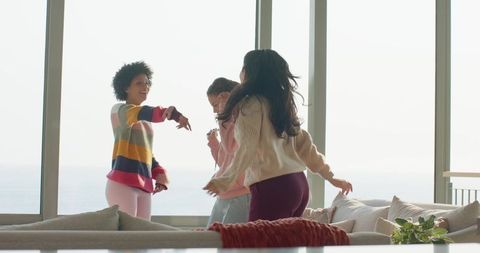 Female friends dancing and laughing on sunlit waterfront sofa with balcony view