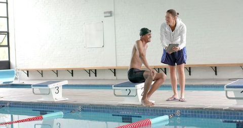 Coach coaching swimmer on starting block using tablet at indoor swimming pool