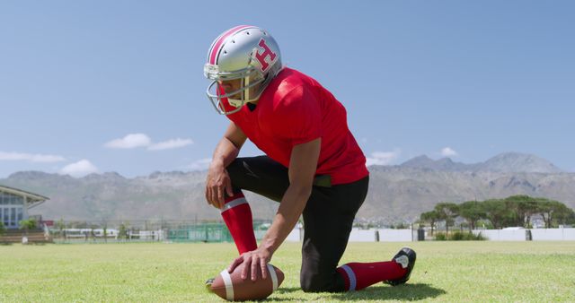 Young American football player kneeling with helmet on field preparing for game. Useful for depicting sportsmanship, teamwork, athletic training, competitive spirit, and physical fitness. Ideal for promoting sports events, youth leagues, athletic gear, and healthy lifestyles.