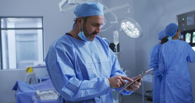 Surgeon standing in operating room interacting with tablet while medical team works in background. Crew wears surgical gowns and caps under bright lights. Useful for content related to advancements in medical technology, modern healthcare practices, or behind-the-scenes looks at surgical procedures.