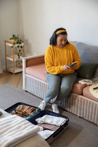 Mature woman sitting on sofa packing suitcase and using smartphone in cozy home for travel
