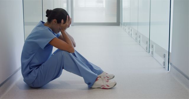 Healthcare worker in blue scrubs is seen sitting on hospital corridor floor with head in hands, indicating stress and exhaustion. This image could be used in articles and discussions about medical professional burnout, workplace stress, healthcare challenges, or hospital environment. It highlights important issues of mental health and job-related fatigue in medical professions.